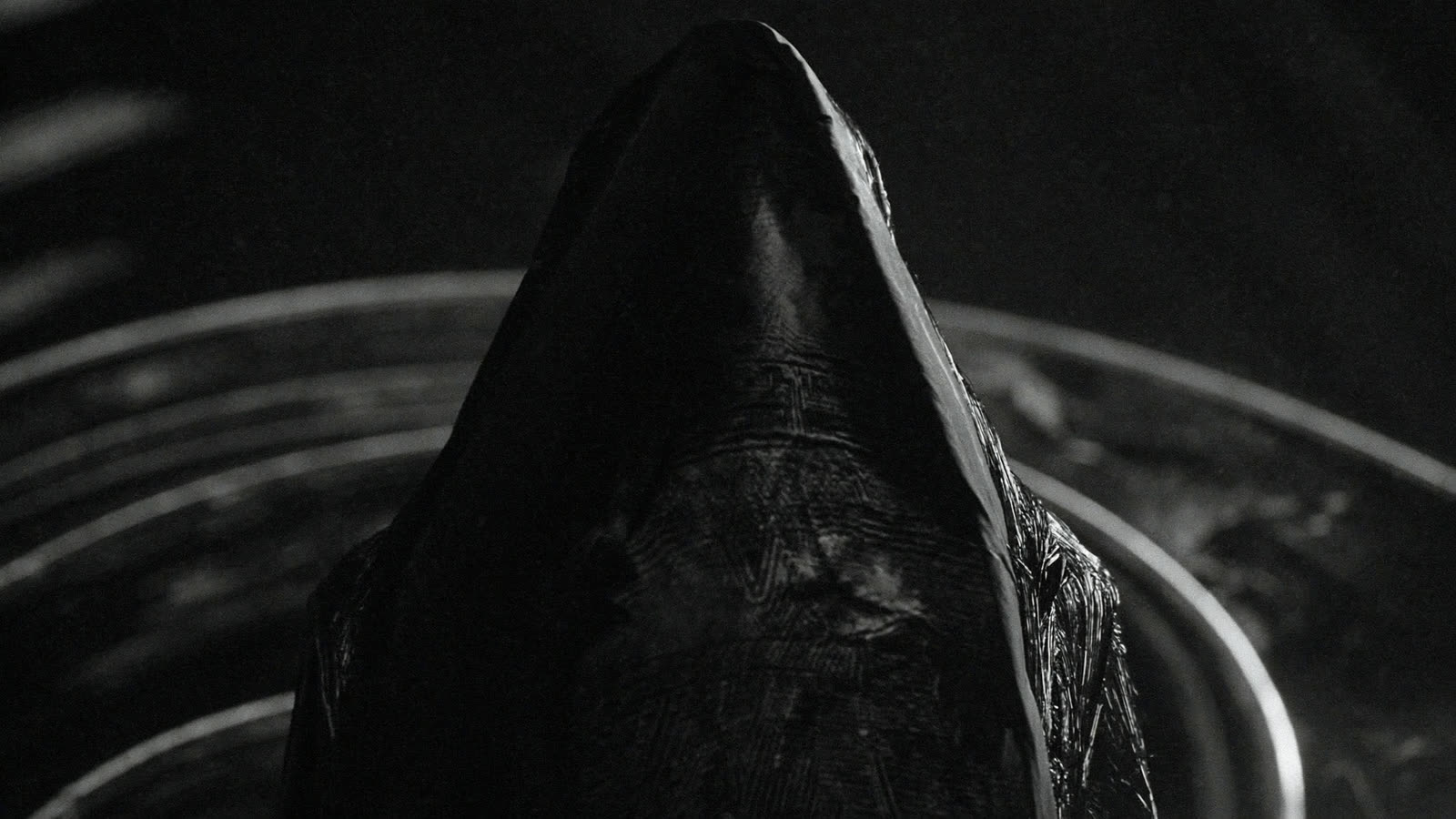 A hooded black figure standing inside concentric metal rings.
